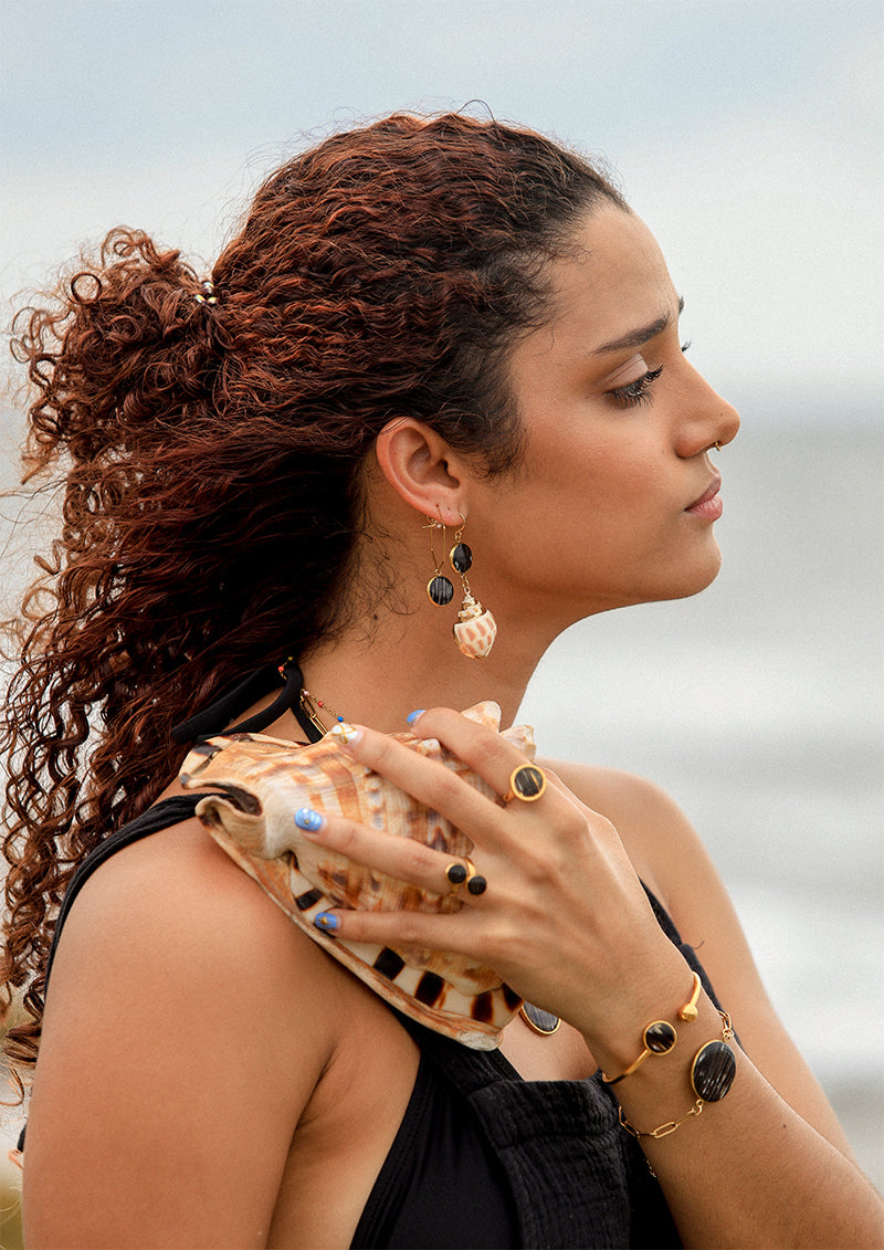 A woman holding a conch shell in a black top wearing two bracelets, two rings, two earrings.
