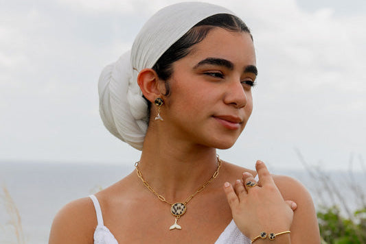 A woman in a white dress and white hair scarf wearing a bracelet, a ring, a necklace and pair of earrings standing in front of a grassy area. 