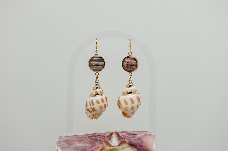 A pair of Leopard Conch Shell Dangles on a white background with a pink shell at the bottom of the image. 