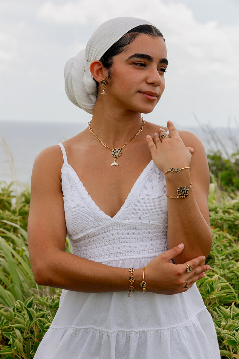 A woman in a white dress and white hair scarf wearing four bracelets, three rings, a necklace and pair of earrings standing in front of a grassy area. 