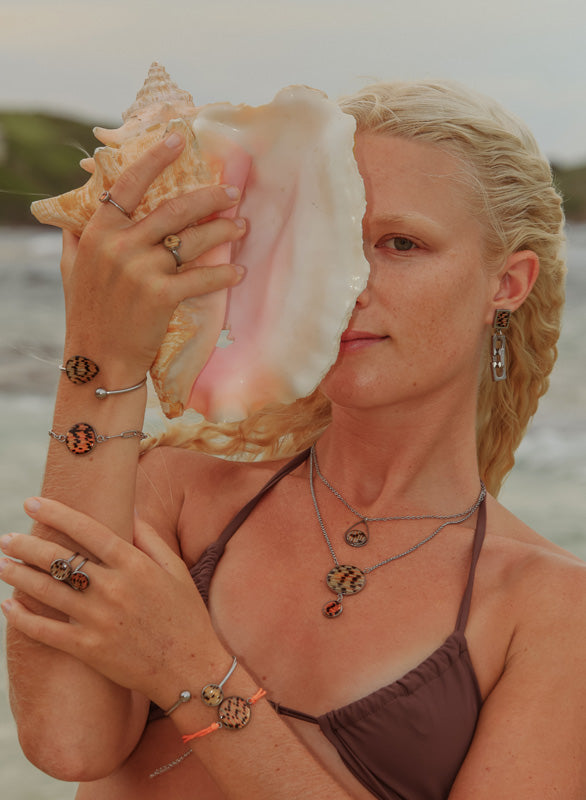 A woman in a brown bikini top in front of the ocean holding a conch shell and wearing three rings, four bracelets, two necklaces and earrings. 