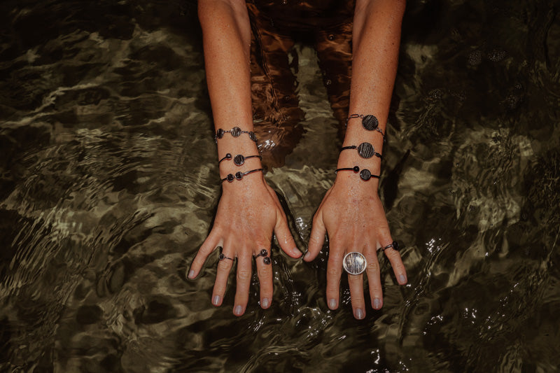 A person's Hands and arms showing four rings, six bracelets over a dark shallow watery background. 