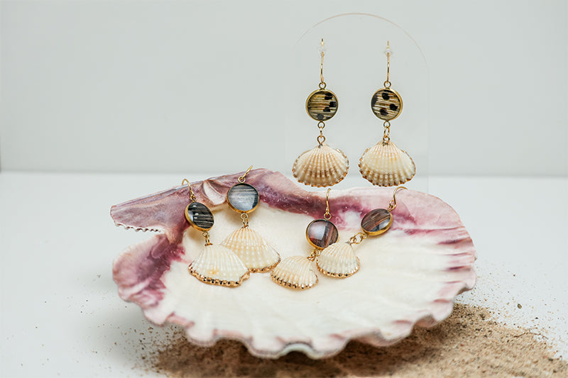 Small Scallop Shell Drops on a stand on a white background with two pairs in a pink clam shell at the bottom of the image with some sand. 
