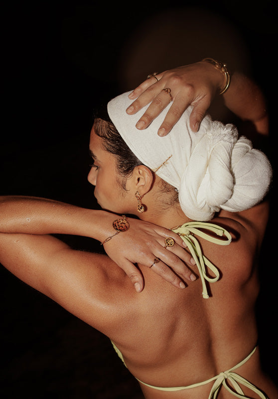 A woman wearing a yellow bikini top, white scarf on her head, two bracelets, two rings, earrings on a dark background. 