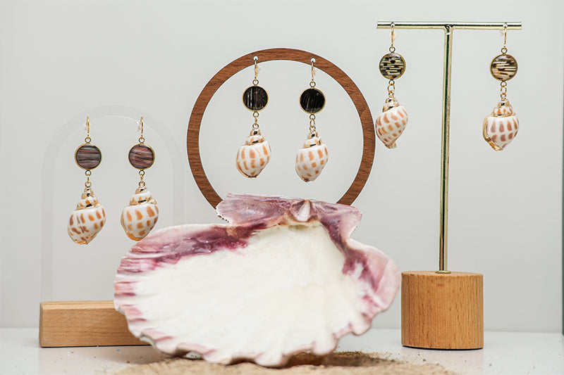 Three pairs of Leopard Conch Shell Dangles on stands on a white background with a pink clam shell at the bottom of the image. 