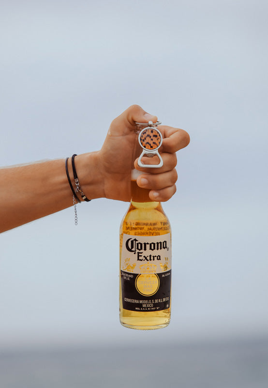 A hand with two bracelets holding a bottle of beer and a Silver Bottle Opener Keychain on a white background.