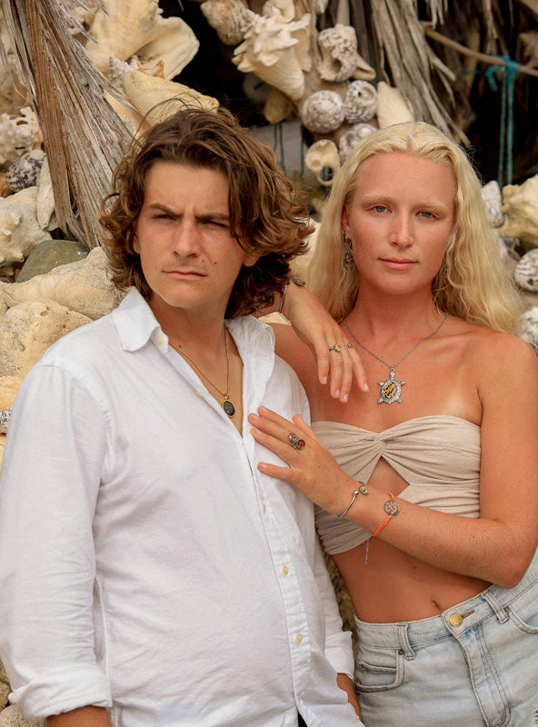 A man in a white shirt wearing. anecklace and a woman in jeans and a cream tube top wearing a necklace, two bracelets, three rings and earrings. They are standing in front of shells and rocks. 