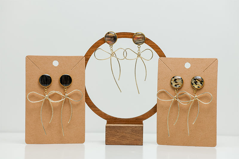 Three pairs of Bows Before Bros Earrings on stands on a white background. 